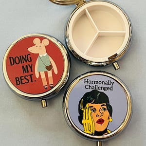 May include: Three round metal pill cases with hinged lids. One case is open, revealing a white plastic interior with three compartments. The other two cases are closed, featuring circular graphics: a mouse with the text DOING MY BEST and a person with the text Hormonally Challenged.
