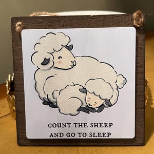 May include: A wooden sign with a white background featuring a cartoon illustration of two sheep. The sign has the text "COUNT THE SHEEP AND GO TO SLEEP" in black. The sign is framed in dark brown and has a jute rope for hanging.