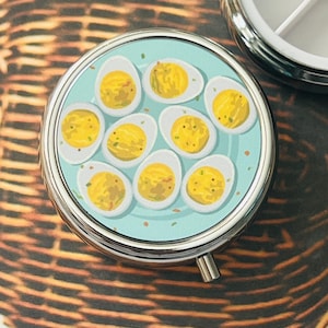 May include: A round, silver-toned metal pill case featuring a top graphic of eight halved deviled eggs with yellow yolks and green garnish on a light blue background. The case has a hinged lid and a small push-button latch for opening and closing.