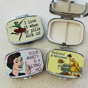 May include: Four silver pill boxes with retro-style illustrations and text. One box is open, revealing two compartments and a mirror. The other boxes feature phrases like "I love it when my pills kick in!" and "Your anxiety is a lying ho."