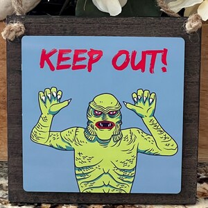 May include: A wooden sign with a light blue background featuring a green creature with the text "KEEP OUT!" in red. The sign is framed in dark brown and has a jute rope for hanging.