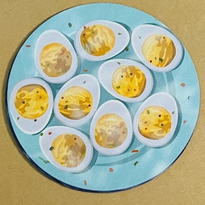 May include: A light blue plate with nine halved deviled eggs. The eggs have yellow yolks with a speckled appearance and are garnished with green herbs. The plate has a slightly textured surface and is set against a tan background.