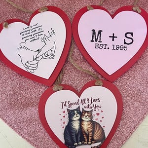 May include: Three heart-shaped ornaments with red borders and twine hangers. One features a quote and names, another has initials and the year 1995, and the third displays a cat illustration with text.
