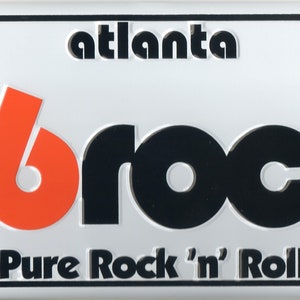 May include: A white metal license plate with black and orange lettering. The text reads "Atlanta 96 Rock Pure Rock 'n' Roll".