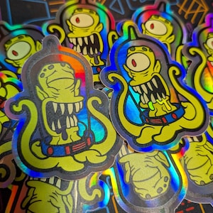 May include: A selection of holographic stickers depicting a cartoon alien with a large mouth, a single eye, and a yellow body. The stickers have a black outline and a blue, green, and orange iridescent effect.