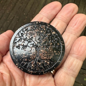 May include: A circular black disc featuring a raised, textured tree of life design in a metallic copper finish. The tree motif covers the entire surface, displaying intricate branches and roots that contrast against the smooth black background.