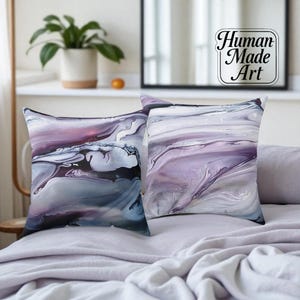 Può includere: Due cuscini quadrati con arte astratta nei toni del viola, grigio e bianco. I cuscini hanno un effetto marmorizzato, con motivi a vortice. I cuscini sono su un letto con una trapunta grigia. I cuscini sono etichettati "Human Made Art".