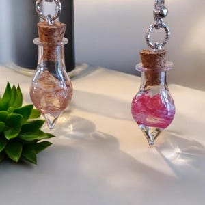 Pode incluir: Dois colares de garrafa de vidro com rolhas de cortiça. Uma garrafa contém cristais rosa e brancos, a outra contém flores rosa e brancas.
