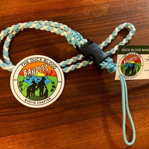 May include: A blue and white paracord lanyard with a round, white sticker featuring the text "The Duck Blood Bandits" and a cartoon image of three people standing in front of a lake. The sticker also includes the text "Est. 2018" and "Martin Chapter". A business card with the text "Duck Blood Bandits Paracord Outfitting" is attached to the lanyard.