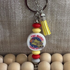 May include: A white charm with the text "The Magic School Bus" and a cartoon image of a yellow school bus flying through space. The charm is attached to a silver key chain with a yellow tassel and a string of colorful wooden beads.