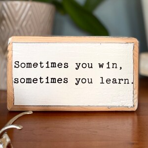 May include: Wooden sign with the text "Sometimes you win, sometimes you learn." The sign is painted white with black lettering and has a light wood frame.