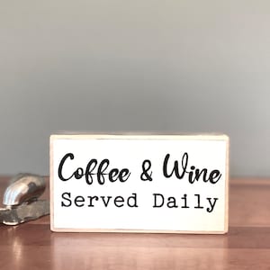 May include: White wooden sign with black text that reads "Coffee & Wine Served Daily". The sign is sitting on a wooden surface.