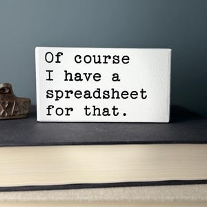 May include: White wooden block with black text that reads "Of course I have a spreadsheet for that."
