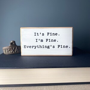 May include: White wooden block sign with the text "It's Fine. I'm Fine. Everything's Fine."