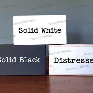 May include: Three wooden blocks with different paint finishes. The top block is white with black text that reads "Solid White". The middle block is black with white text that reads "Solid Black". The bottom block is white with black text that reads "Distressed".