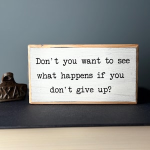 Puede incluir: Un letrero de madera blanco con el texto "Don't you want to see what happens if you don't give up?" en fuente de m&aacute;quina de escribir negra.
