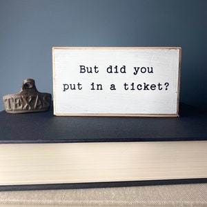 May include: White wooden sign with black text that reads "But did you put in a ticket?" The sign is sitting on top of a stack of books.