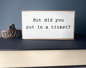 May include: White wooden sign with black text that reads "But did you put in a ticket?" The sign is sitting on top of a stack of books.
