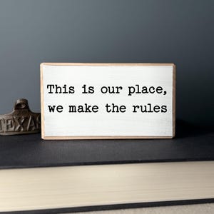May include: A rectangular wooden sign with the text "This is our place, we make the rules" in black font. The sign has a white painted surface and a natural wood border. The sign is on top of a stack of books.