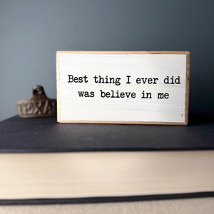 May include: White wooden sign with the text "Best thing I ever did was believe in me" in black typewriter font.