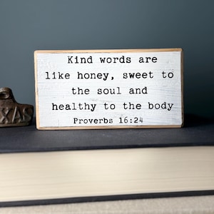 May include: White wooden sign with black text that reads "Kind words are like honey, sweet to the soul and healthy to the body Proverbs 16:24".