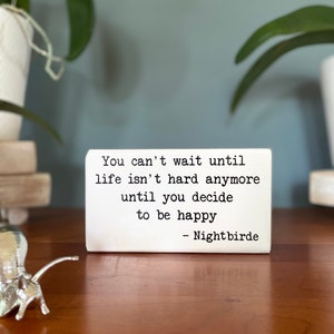May include: White wooden sign with black text that reads "You can't wait until life isn't hard anymore until you decide to be happy - Nightbirde".