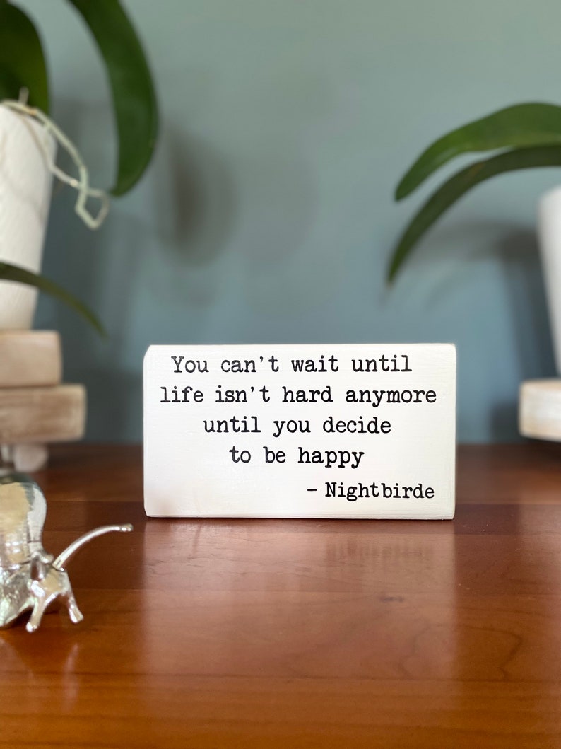 May include: White wooden sign with black text that reads "You can't wait until life isn't hard anymore until you decide to be happy - Nightbirde".