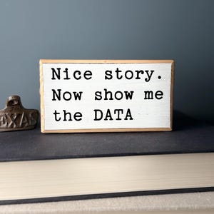 May include: White wooden sign with black text that reads "Nice story. Now show me the DATA".
