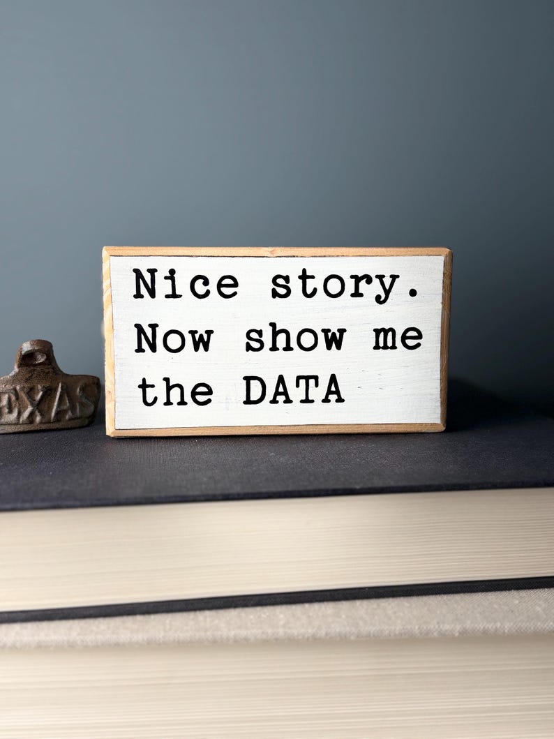 May include: White wooden sign with black text that reads "Nice story. Now show me the DATA".