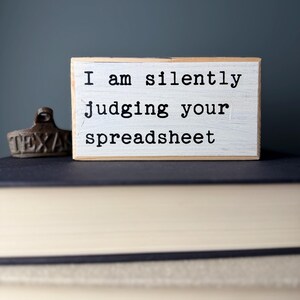 May include: White wooden sign with black text that reads "I am silently judging your spreadsheet".