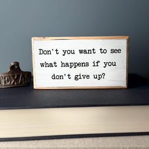Puede incluir: Un letrero de madera blanco con texto negro que dice "Don't you want to see what happens if you don't give up?"