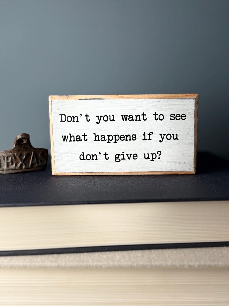 Puede incluir: Un letrero de madera blanco con texto negro que dice "Don't you want to see what happens if you don't give up?"
