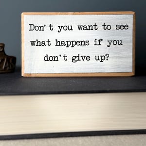 Puede incluir: Bloque de madera blanco con el texto "Don't you want to see what happens if you don't give up?" impreso en negro.