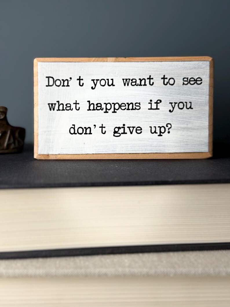 Puede incluir: Bloque de madera blanco con el texto "Don't you want to see what happens if you don't give up?" impreso en negro.