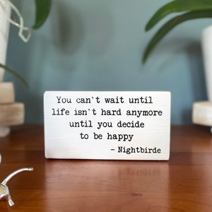 May include: White wooden sign with black text that reads "You can't wait until life isn't hard anymore until you decide to be happy - Nightbirde".