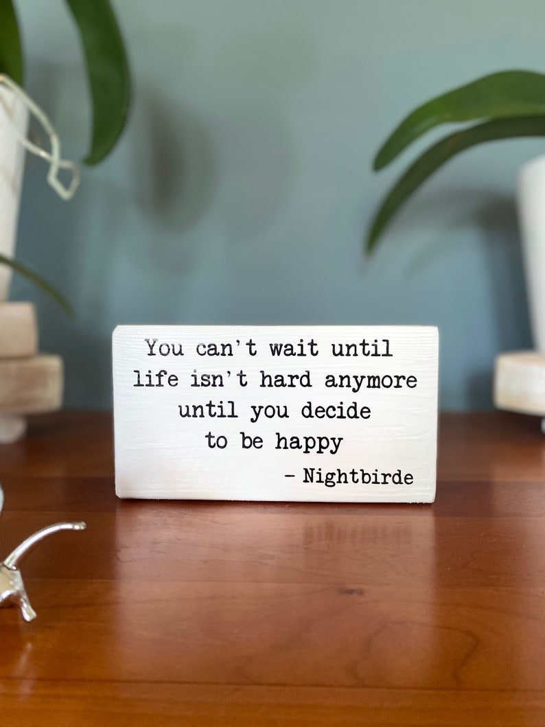 May include: White wooden sign with black text that reads "You can't wait until life isn't hard anymore until you decide to be happy - Nightbirde".