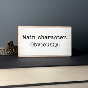 May include: A small, rectangular wooden sign with the text "Main character. Obviously." in black. The sign has a white painted surface and a natural wood frame. A small, brown metal object with the word "TEXAS" is to the left of the sign.