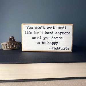 May include: A white wooden sign with the text "You can't wait until life isn't hard anymore until you decide to be happy - Nightbirde".