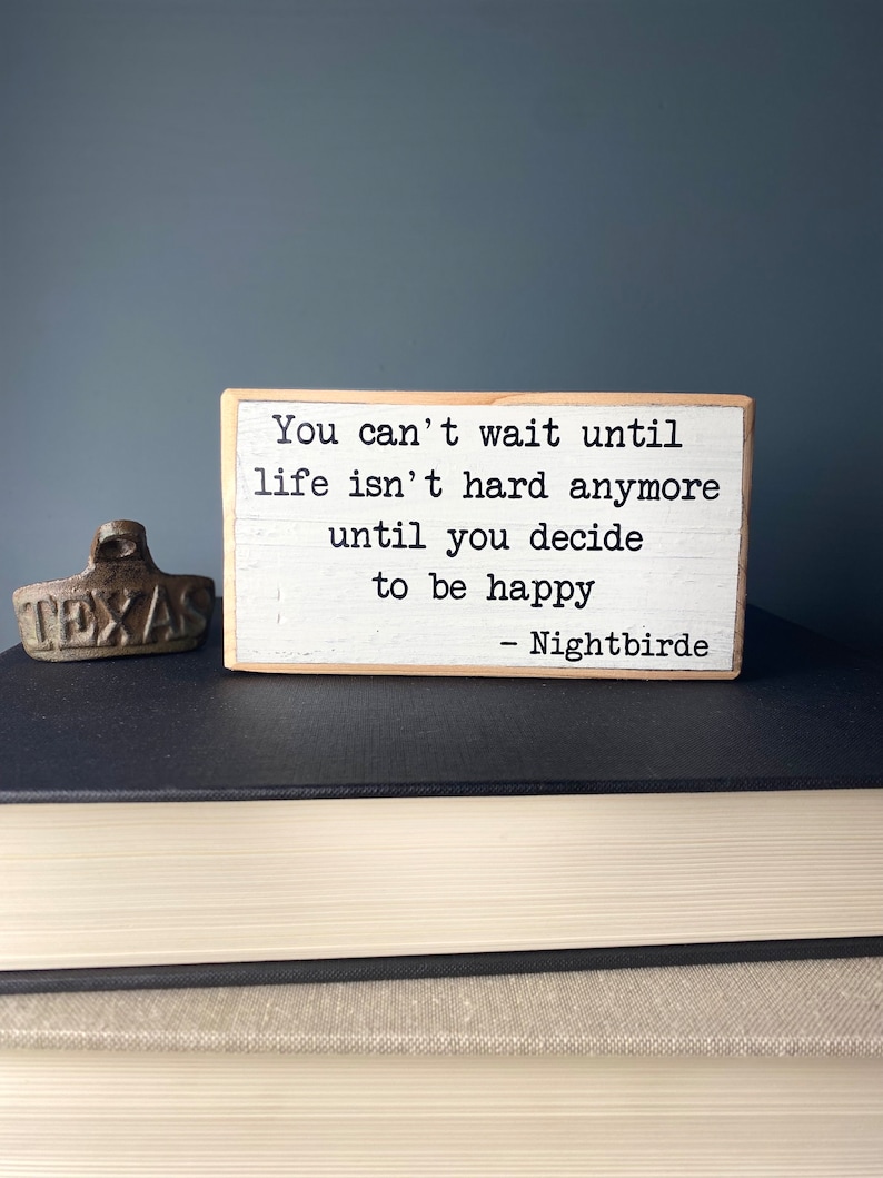 May include: A white wooden sign with the text "You can't wait until life isn't hard anymore until you decide to be happy - Nightbirde".