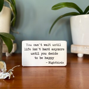 May include: White wooden block with a black and white inspirational quote that reads "You can't wait until life isn't hard anymore until you decide to be happy - Nightbirde"
