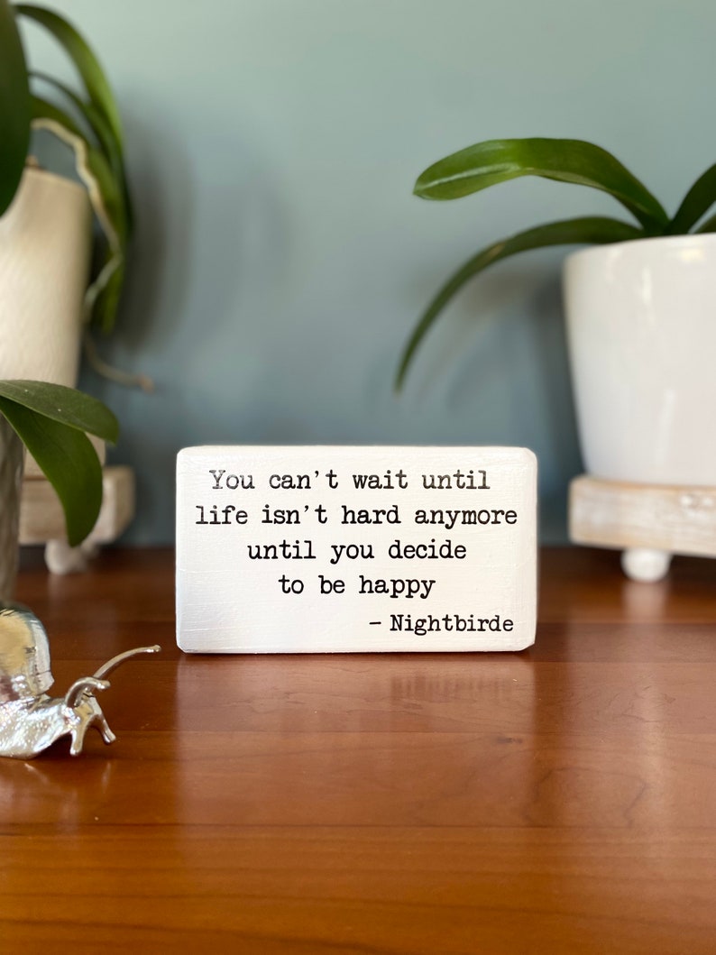May include: White wooden block with a black and white inspirational quote that reads "You can't wait until life isn't hard anymore until you decide to be happy - Nightbirde"