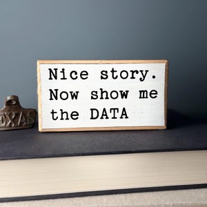 May include: White wooden block with the text "Nice story. Now show me the DATA" in black lettering.