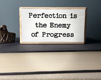 May include: White wooden block sign with black text that reads "Perfection is the Enemy of Progress".