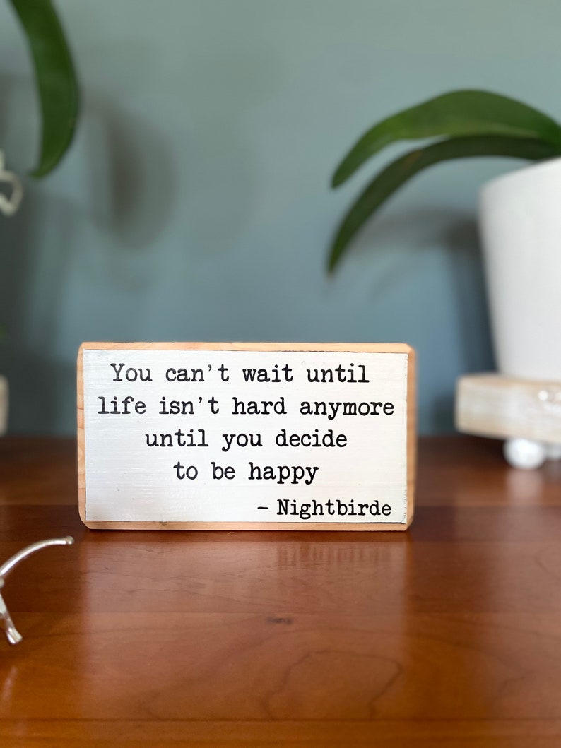 May include: A wooden sign with a white background and black text. The text reads: "You can't wait until life isn't hard anymore until you decide to be happy - Nightbirde"