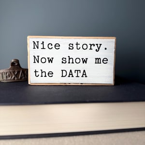 May include: A white wooden sign with black text that reads "Nice story. Now show me the DATA".