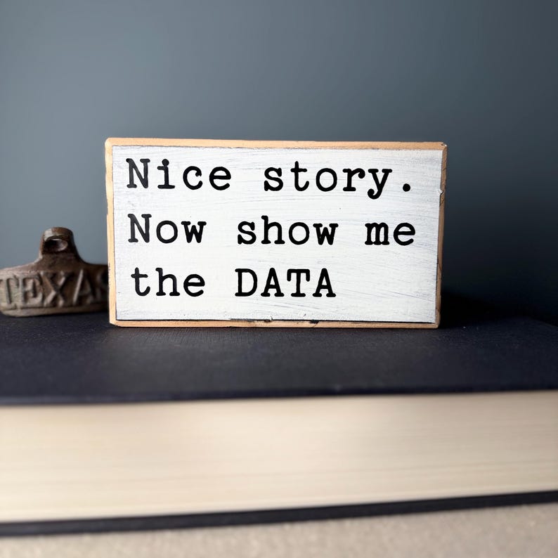 May include: A white wooden sign with black text that reads "Nice story. Now show me the DATA".