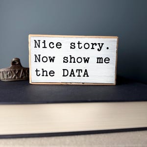 May include: A white wooden sign with black text that reads "Nice story. Now show me the DATA". The sign is sitting on top of a stack of books.