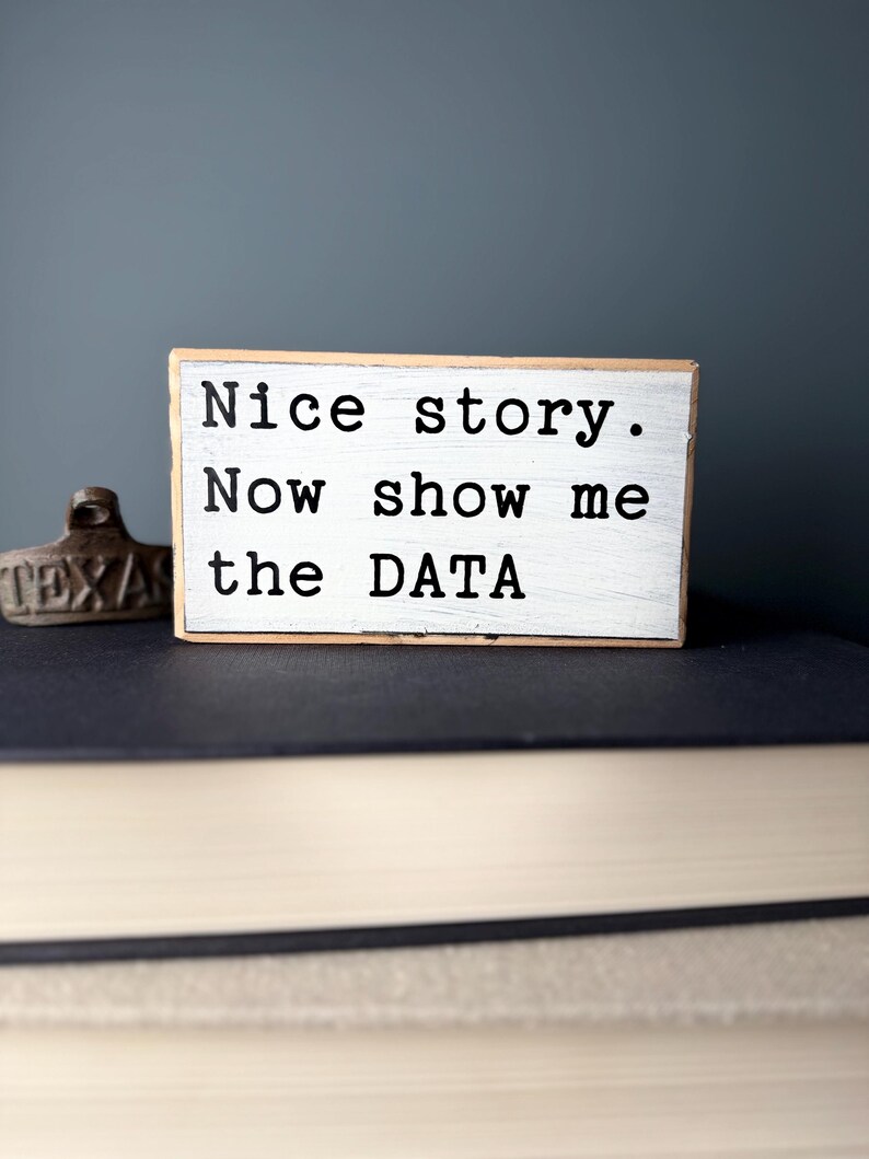 May include: A white wooden sign with black text that reads "Nice story. Now show me the DATA". The sign is sitting on top of a stack of books.