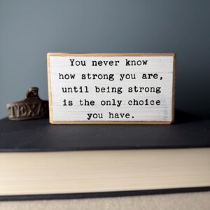 May include: A wooden sign with a white background and black text that reads "You never know how strong you are, until being strong is the only choice you have."