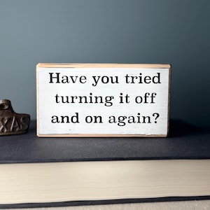 May include: A small wooden sign with white paint and black lettering. The sign reads "Have you tried turning it off and on again?"